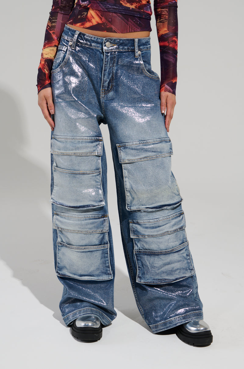 A person wearing the TO THE MOON DENIM PANT featuring a shiny fabric, wide-leg silhouette, and large pockets, paired with a long-sleeve patterned top and black shoes.