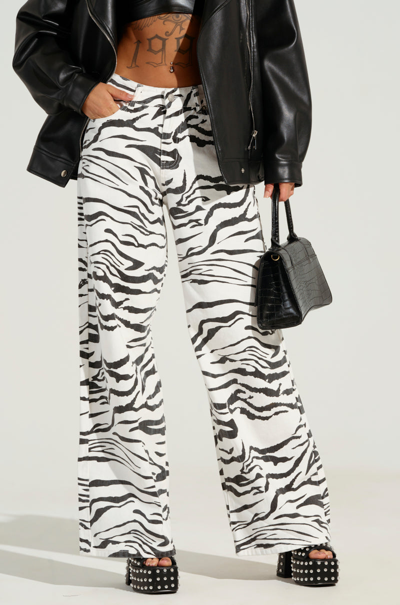 Someone dressed in a black leather jacket, BACK IT UP ZEBRA PANT showcasing a wide leg silhouette, and studded platform shoes, holding a small black handbag. A tattoo can be partially seen beneath the jacket.