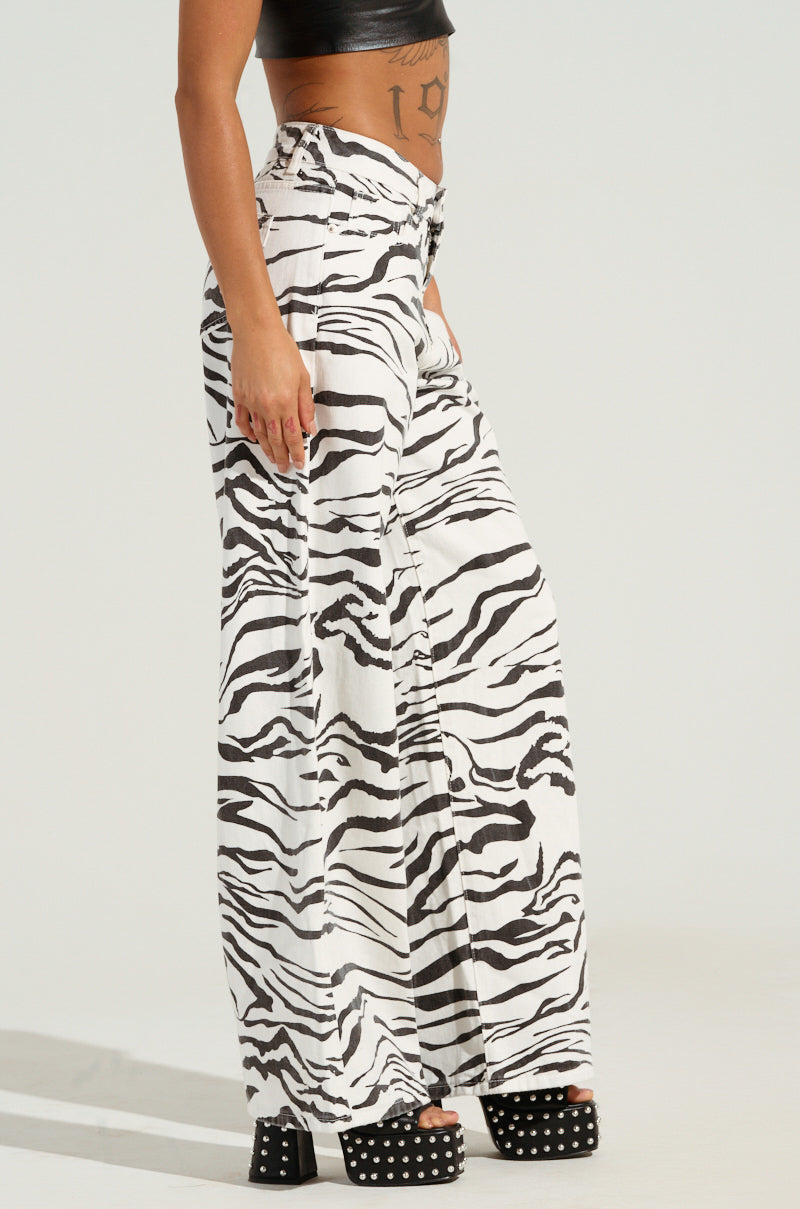 A person is wearing high-rise BACK IT UP ZEBRA PANTs, paired with a black crop top and black platform sandals featuring silver studs. The plain background accentuates the striking zebra-patterned ensemble.
