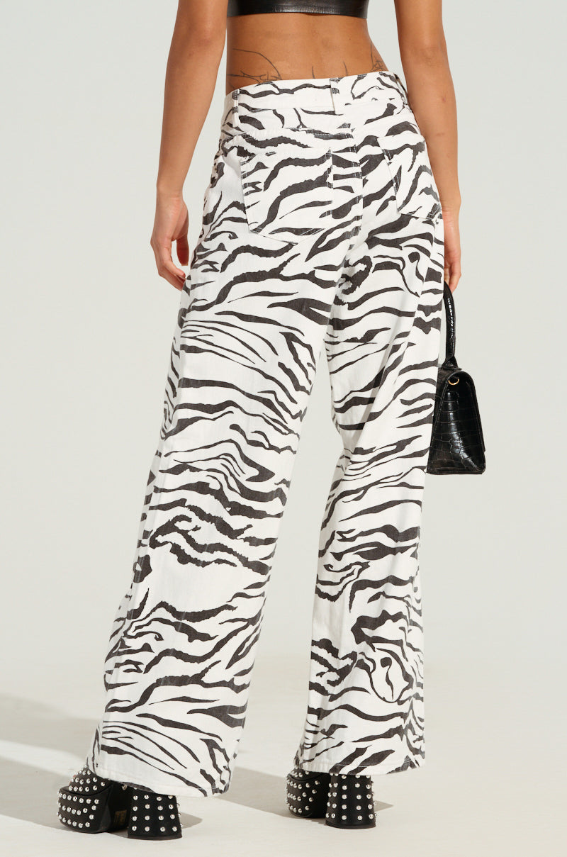 The individual is dressed in BACK IT UP ZEBRA PANTS, paired with a black crop top and studded black high heels. They are carrying a black handbag, with the plain background highlighting the striking zebra pants and stylish outfit.
