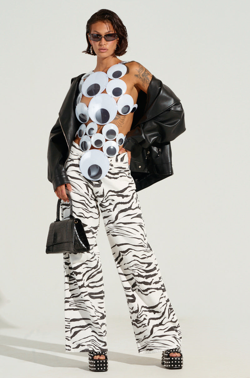 A person confidently stands against a plain background, sporting a unique outfit featuring large circular cutouts on the top, BACK IT UP ZEBRA PANT with a high-rise and wide-leg silhouette, a black leather jacket, sunglasses, and holding a small black handbag.