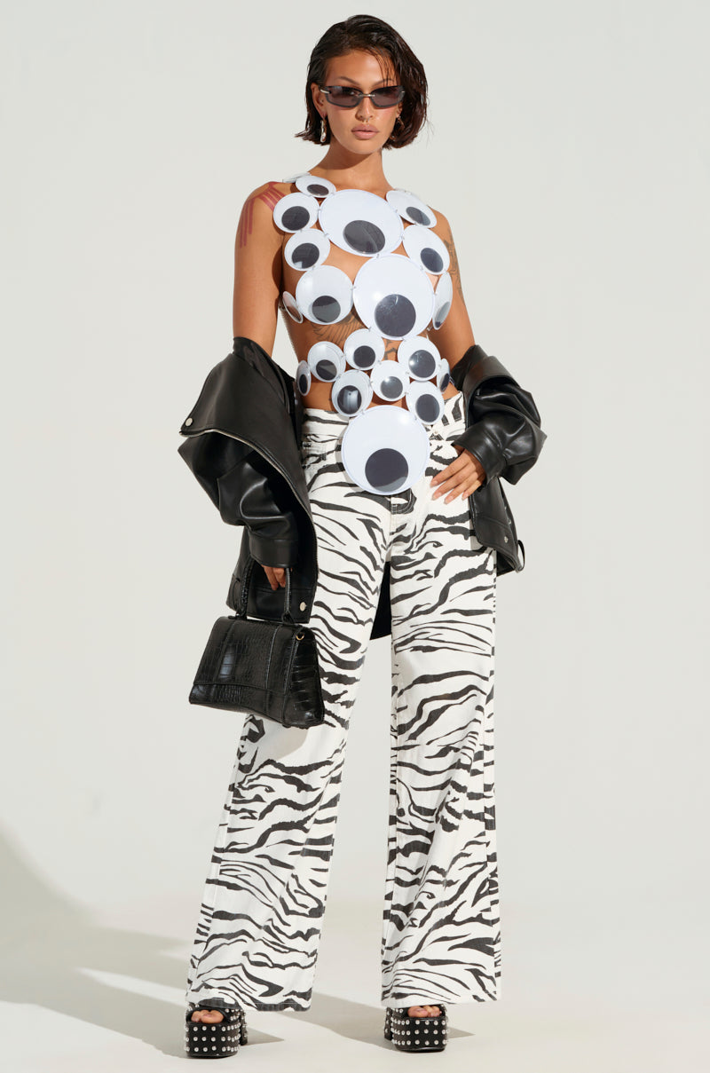 A person dressed in a fashion-forward ensemble featuring an artistic top with large circular patterns, the BACK IT UP ZEBRA PANT in a high-rise wide-leg silhouette, and platform shoes. They hold a black jacket and a purse, wearing sunglasses against a plain background.