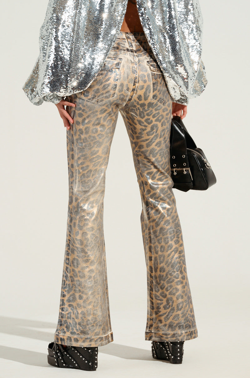 A person dressed in the FIERCE CHEETAH PANT, a silver sequined jacket, and black studded platform shoes strikes a pose with a flared leg silhouette, holding a black handbag against a light background.