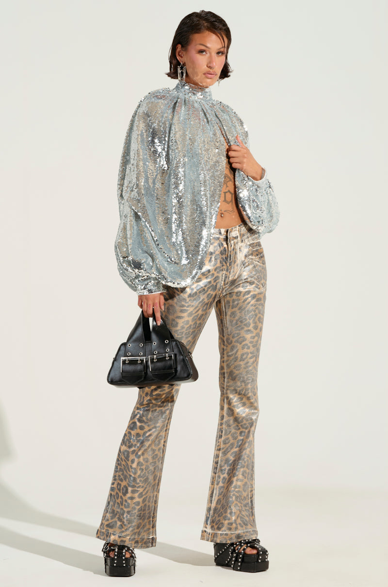 A person confidently poses in a dazzling silver sequined top and the FIERCE CHEETAH PANT, featuring a flared leg silhouette. They carry a black handbag and wear platform sandals, while the plain background accentuates their strikingly stylish ensemble.