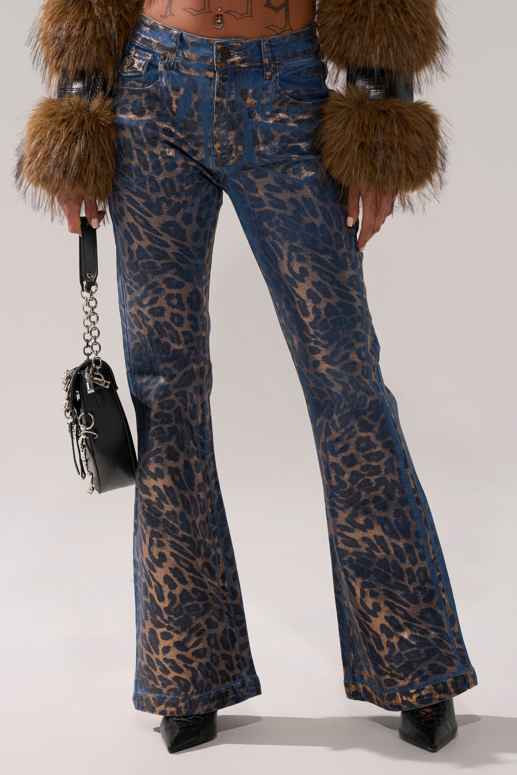 Wearing CHEETAH GIRLS FLARE DENIM blue jeans with bold metallic cheetah print, a brown fur-trimmed jacket, and black pointed-toe shoes, a person holds a small black handbag with silver chains.