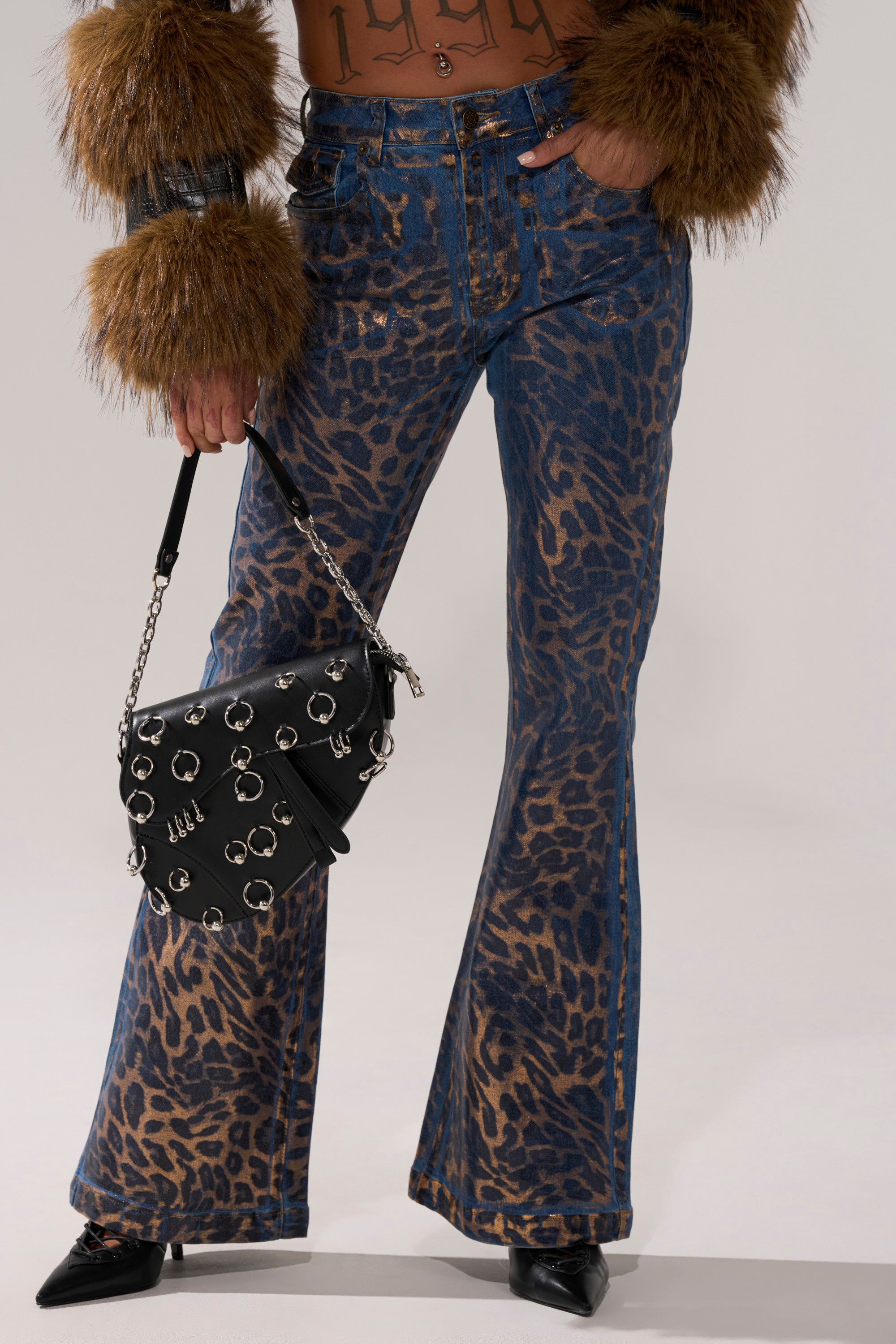 Wearing CHEETAH GIRLS FLARE DENIM jeans in metallic cheetah print, a sheer brown top with faux fur cuffs, and pointed black shoes, the person holds a black handbag decorated with metal rings and embellishments.