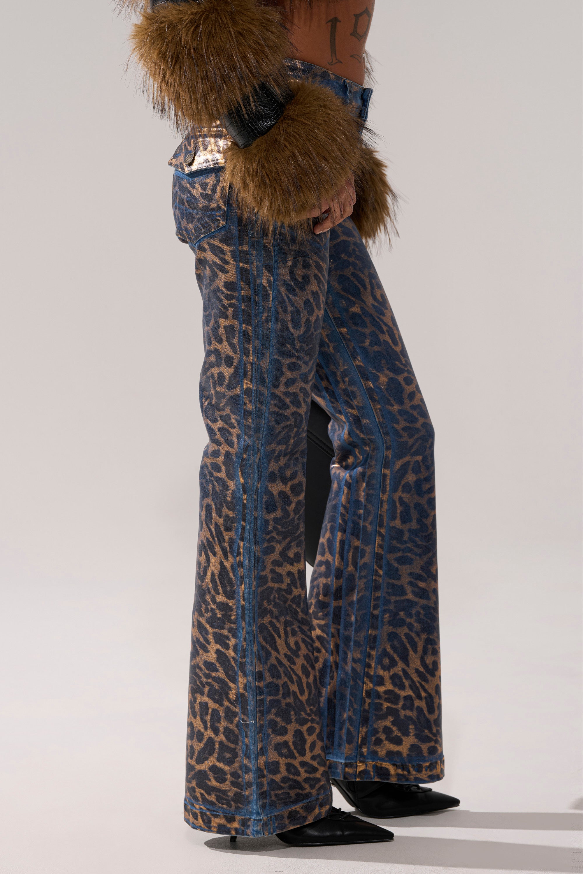 Wearing the CHEETAH GIRLS FLARE DENIM in metallic cheetah print, paired with black pointed shoes and furry brown sleeves, a person is shown from the torso down against a plain background.