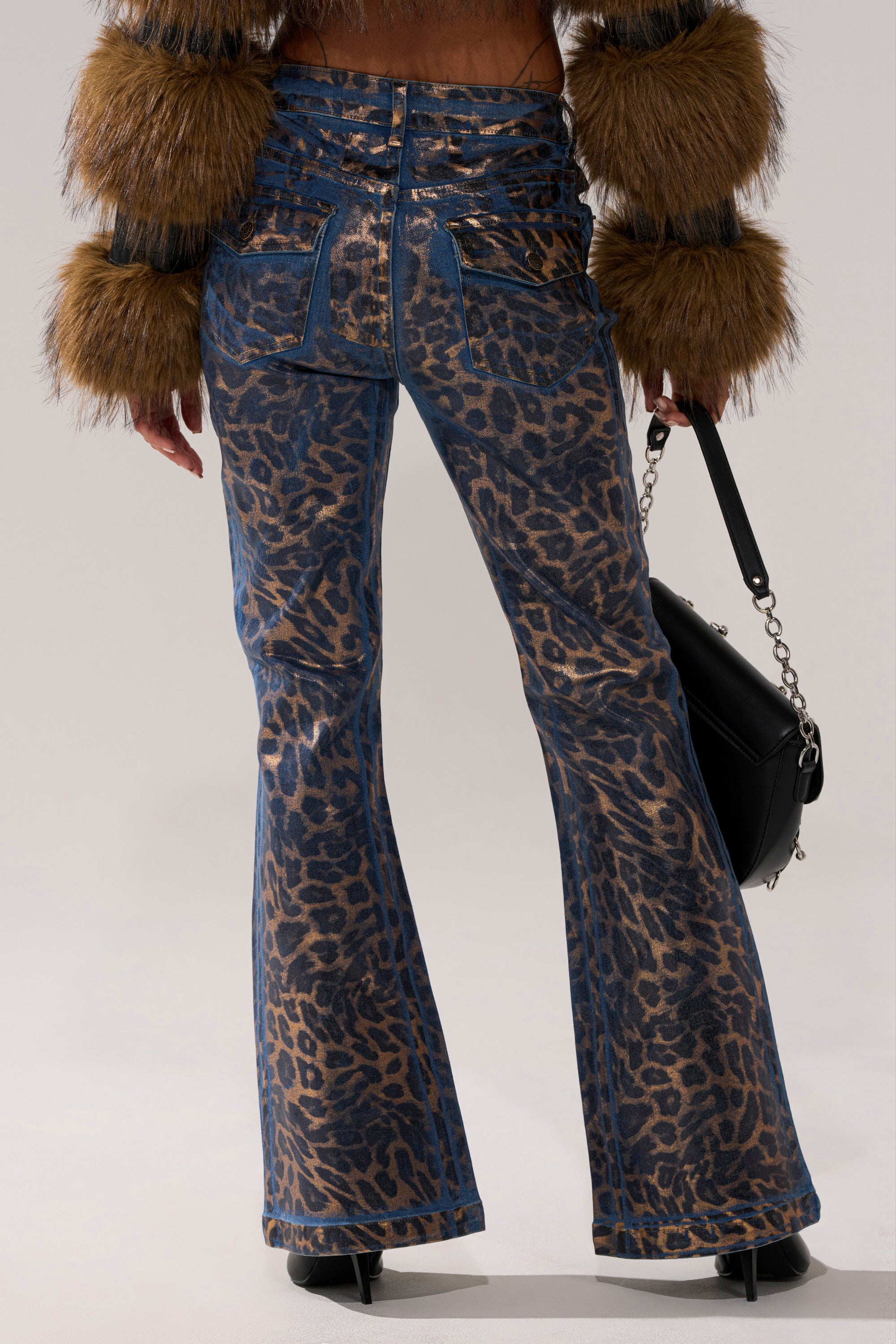 A person stands facing away, wearing CHEETAH GIRLS FLARE DENIM—mid-rise, flared-leg jeans in leopard print—with a brown faux fur sleeve jacket, black heeled boots, and holding a black chain-strap handbag.