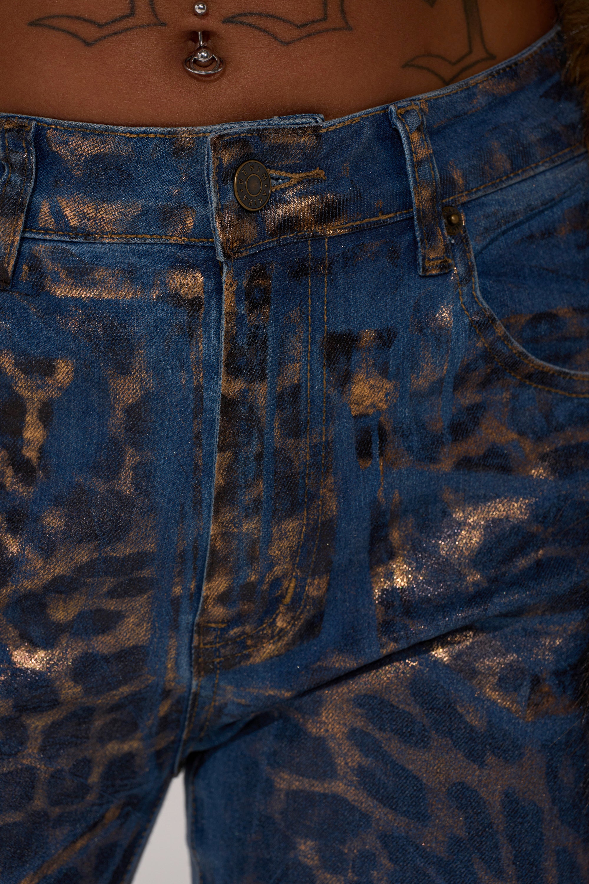 Close-up of the CHEETAH GIRLS FLARE DENIM: blue metallic cheetah print flared-leg jeans, mid-rise fit, shown with a glimpse of a tattooed torso and belly button piercing above the waistband.