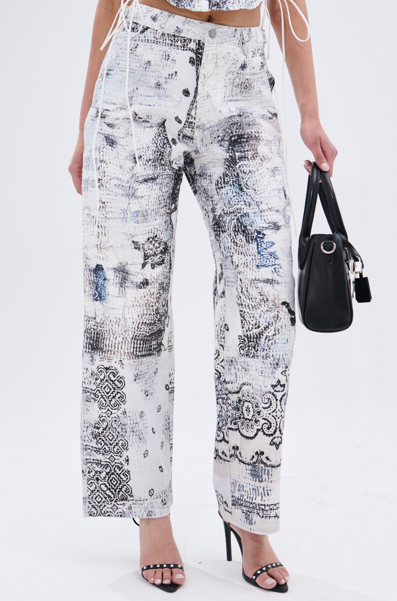 A person styles the AKIRA Label READ ME TROUSER—high-waisted, wide-leg pants with an abstract linework print—with black heels, a matching cropped top, and a black handbag for a chic look.