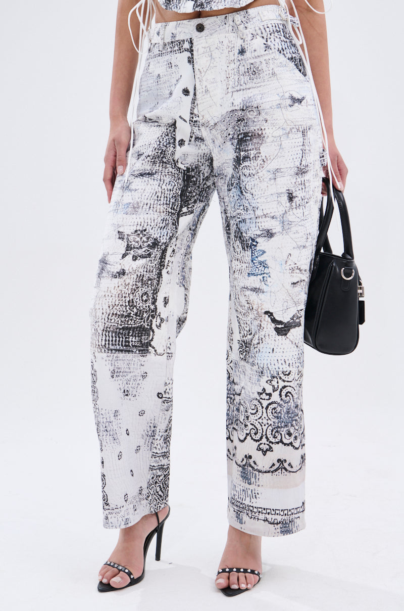 A person wears the AKIRA Label READ ME TROUSER—high-waisted, wide-leg white pants with abstract linework—paired with a matching crop top, black heeled sandals, and a black handbag. Background is plain white.