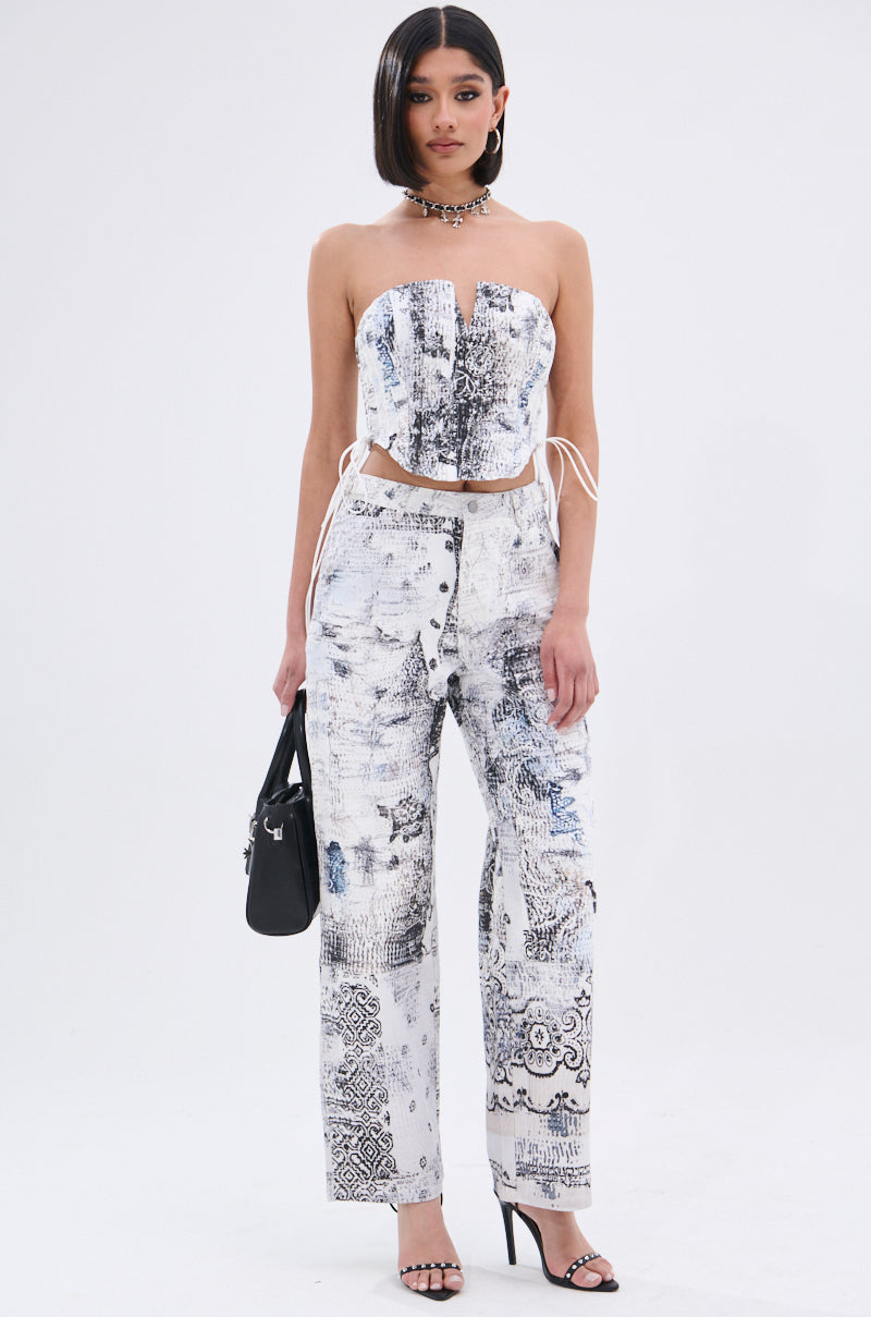 A woman stands against a white background wearing an AKIRA Label strapless top and the READ ME TROUSER featuring an abstract linework print. She holds a black handbag, wears black high-heeled sandals, and accessorizes with a necklace.