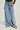 A person wearing the DREAMER WIDE LEG JEAN in light wash denim, paired with a dark green long-sleeved top, is standing against a light background. The high-rise waist jeans have a relaxed fit, and the ensemble is completed with beige shoes featuring shiny details.