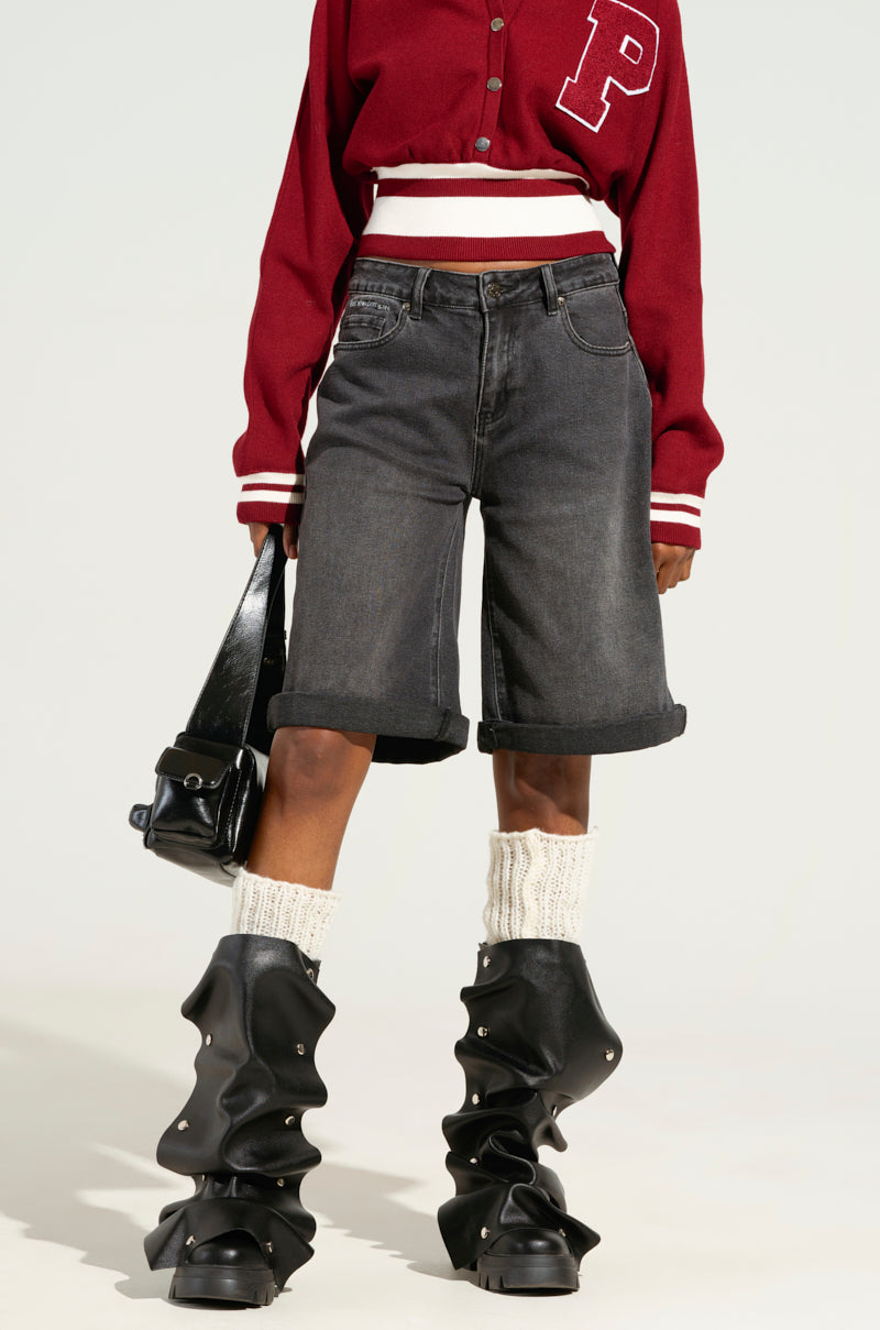 A person is wearing a red varsity jacket featuring a white "P" on the chest, paired with the DONT KNOW DONT CARE BERMUDA SHORT in stretchy black denim, and slouchy black boots. They are carrying a black handbag over their shoulder, with cream knitted socks peeking out from the boots.