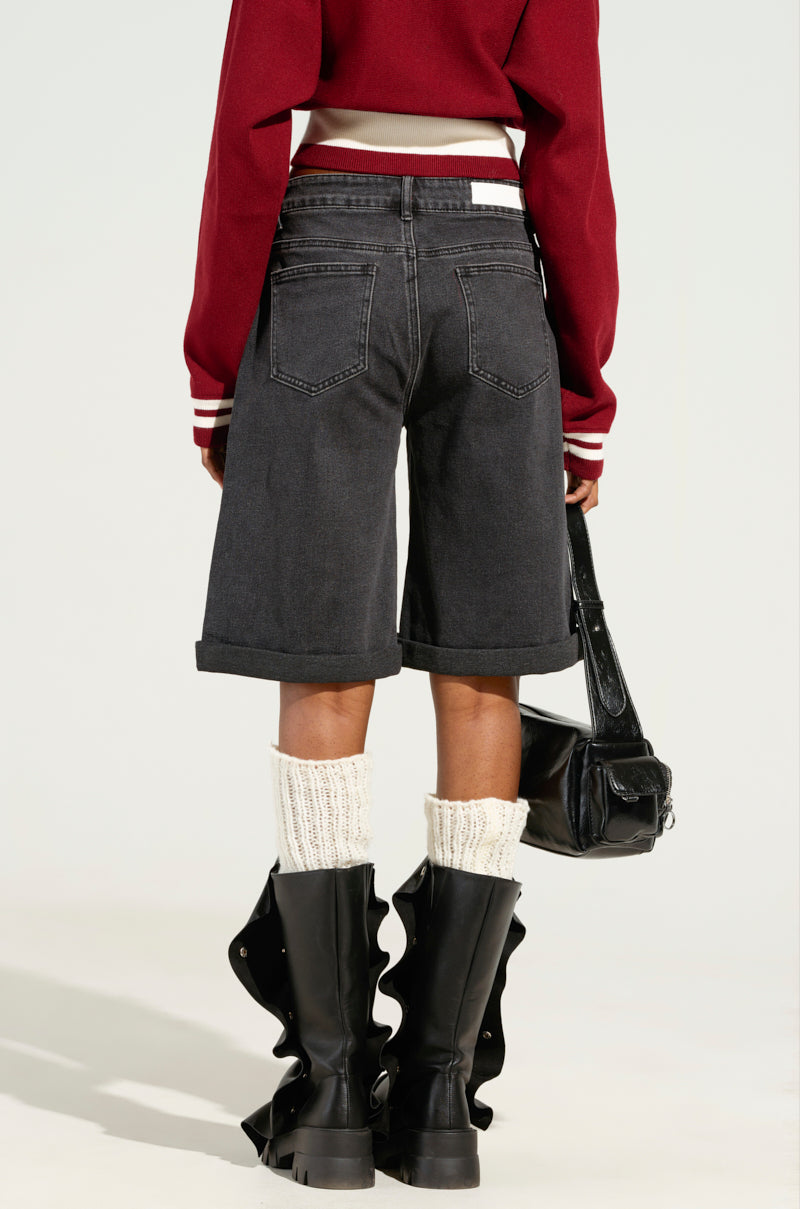 Wearing a striking red jacket and DONT KNOW DONT CARE BERMUDA SHORTS in a dark hue with rolled-up cuffs, the individual is captured from behind. They are adorned with knee-high white socks and black chunky boots featuring ruffle details, and they carry a chic black handbag against a simple backdrop.