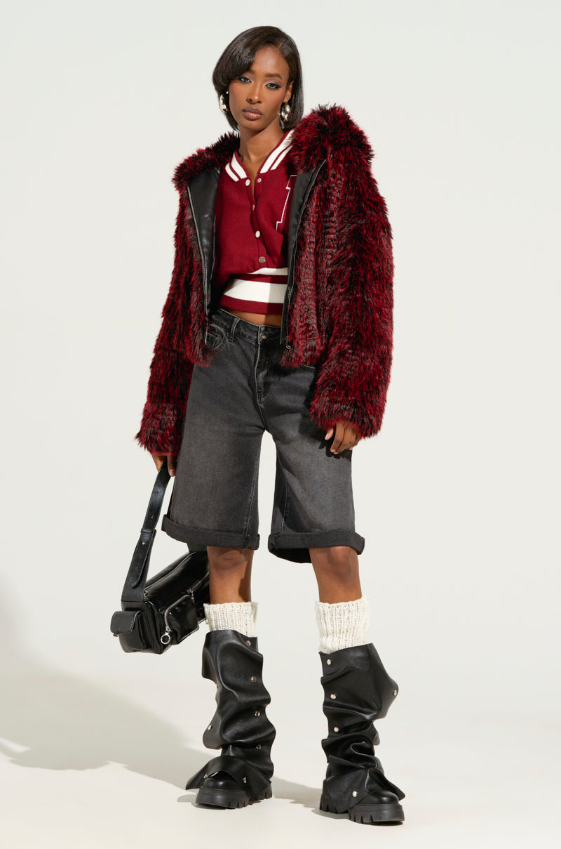 A person confidently stands out in a red faux fur jacket layered over a matching top. They have paired it with stretchy black denim DONT KNOW DONT CARE BERMUDA SHORTS, chunky boots adorned with white socks, and are holding a black bag. The simple background accentuates the distinctive fashion ensemble.