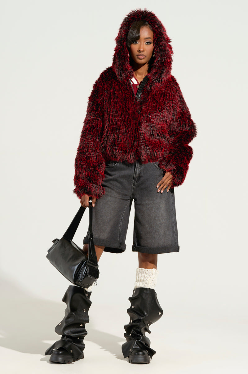 A person stands confidently in a red, plush hoodie paired with DONT KNOW DONT CARE BERMUDA SHORT and unique black boots. They carry a black bag, their dark hair peeking out from the hood. The background is a plain light color, enhancing the casual flair of their wide-leg fit attire.