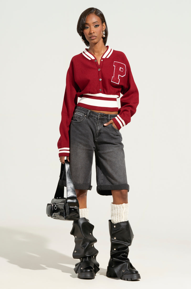 A person poses confidently in a red varsity jacket featuring a prominent "P," paired with the DONT KNOW DONT CARE BERMUDA SHORT in gray and black ruffled boots. They hold a black handbag, with socks visible above their boots, set against a plain background.