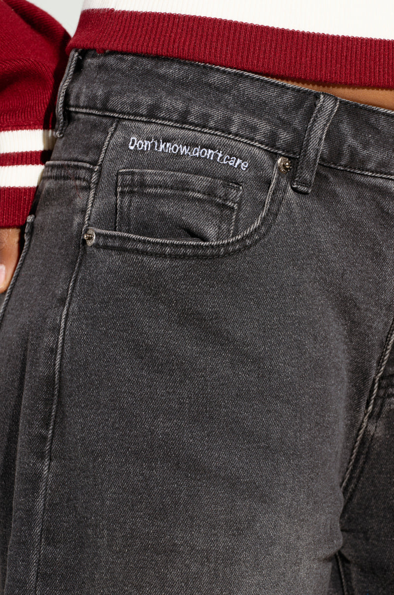 Close-up of a person wearing the DONT KNOW DONT CARE BERMUDA SHORT, featuring stretchy black denim and the phrase "Don't know, don't care" embroidered above the front pocket. The person is also wearing a red and white jacket.