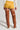 A person stands with their back turned, showcasing the mustard yellow SWEET AS CANDY SHORT in a mini length. They have paired it with a white knitted sweater and tall brown thigh-high leather boots. The simple, minimalist background perfectly emphasizes this chic faux suede outfit.