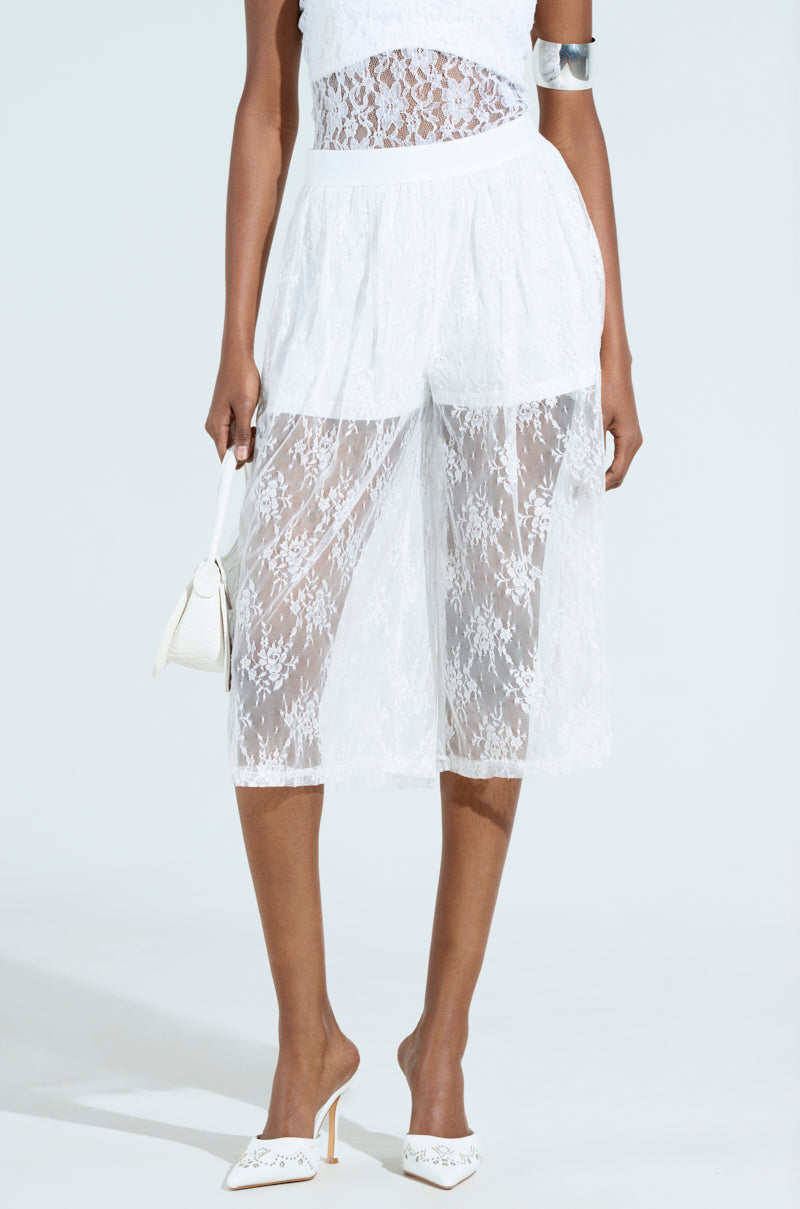 Dressed in the KELLY LACE SHORT outfit with a strapless top and knee-length skirt, a person holds a small white bag, dons an arm cuff, and wears white pointed-toe heeled mules against a light background.