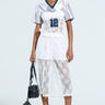 A person in a sporty white top with number 12 over a KELLY LACE SHORT dress, wearing sunglasses, black and white lace-up boots, and holding a black handbag against a plain light-colored backdrop.