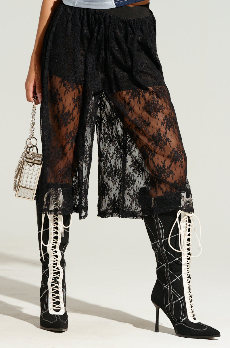 A person wearing the KELLY LACE SHORT IN BLACK with sheer lace pants, paired with black knee-high boots featuring white laces, holds a small cage-like handbag. The ensemble is stylish and fashion-forward.