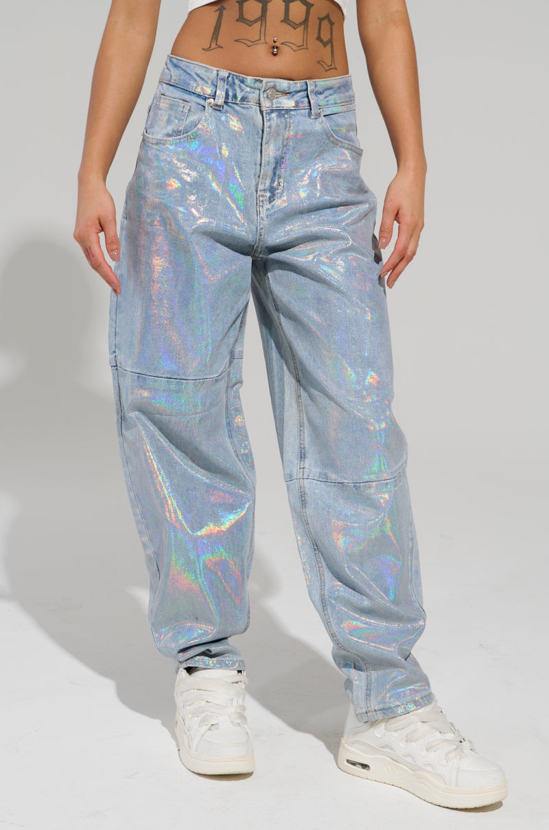 A person wearing the high-rise ASTRONOMICAL Silver Gilded Barrel Denim Pant and white sneakers stands against a plain backdrop. Their midriff is visible, revealing a "1999" tattoo, while the shimmering finish of the denim catches every angle.