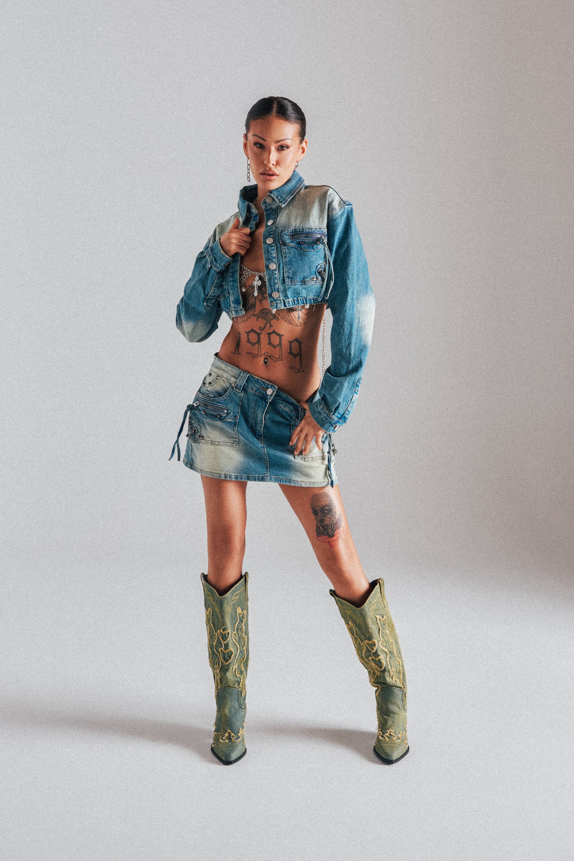 A woman stands confidently in the CROWN CROPPED LONG SLEEVE DENIM BUTTON DOWN, paired with a denim mini skirt and green cowboy boots. Tattoos accent her look, and her hair is tied back against a plain light background.