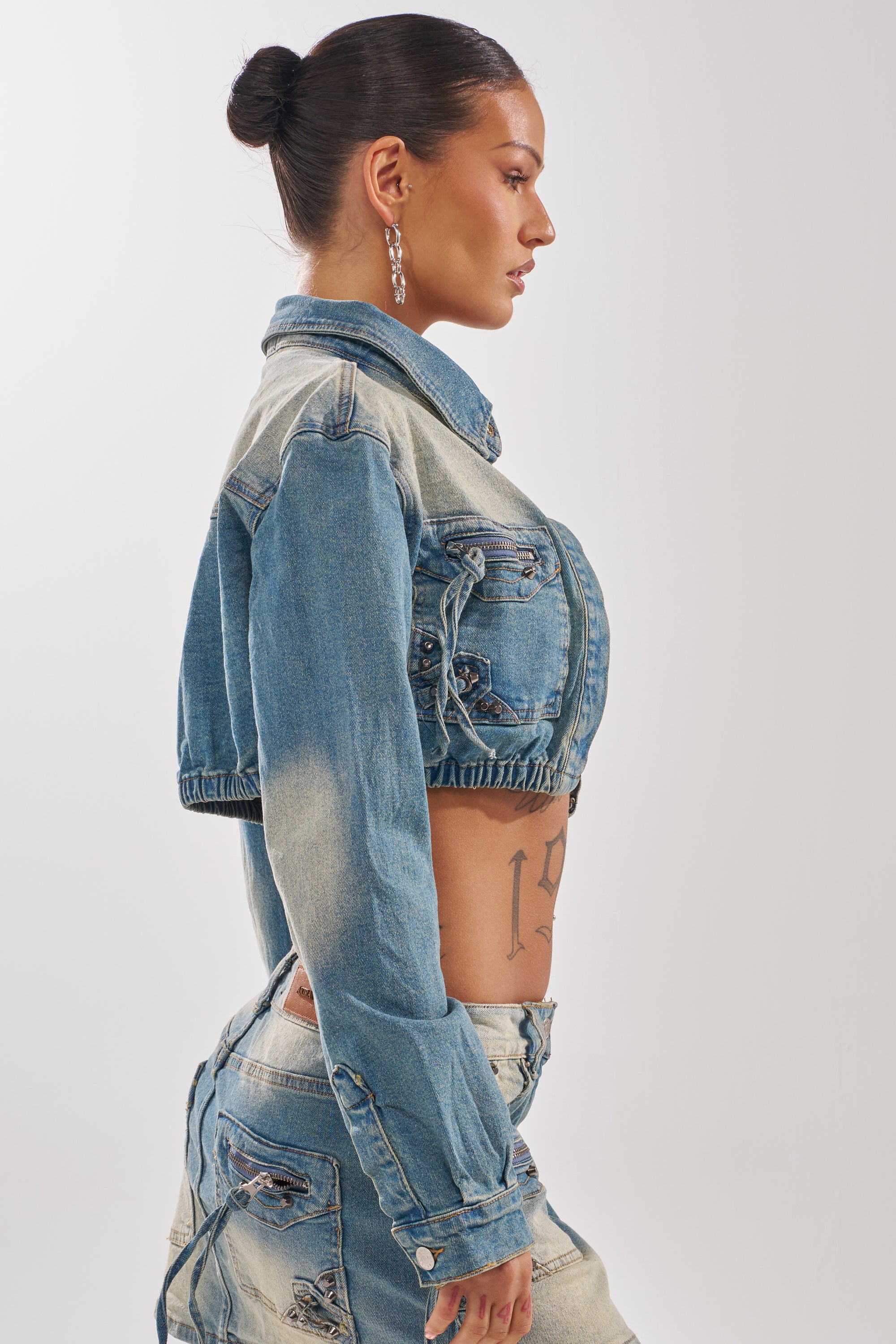 A woman with dark hair in a bun wears the CROWN CROPPED LONG SLEEVE DENIM BUTTON DOWN and matching jeans, seen in profile against a plain light background, revealing a tattoo on her left side.