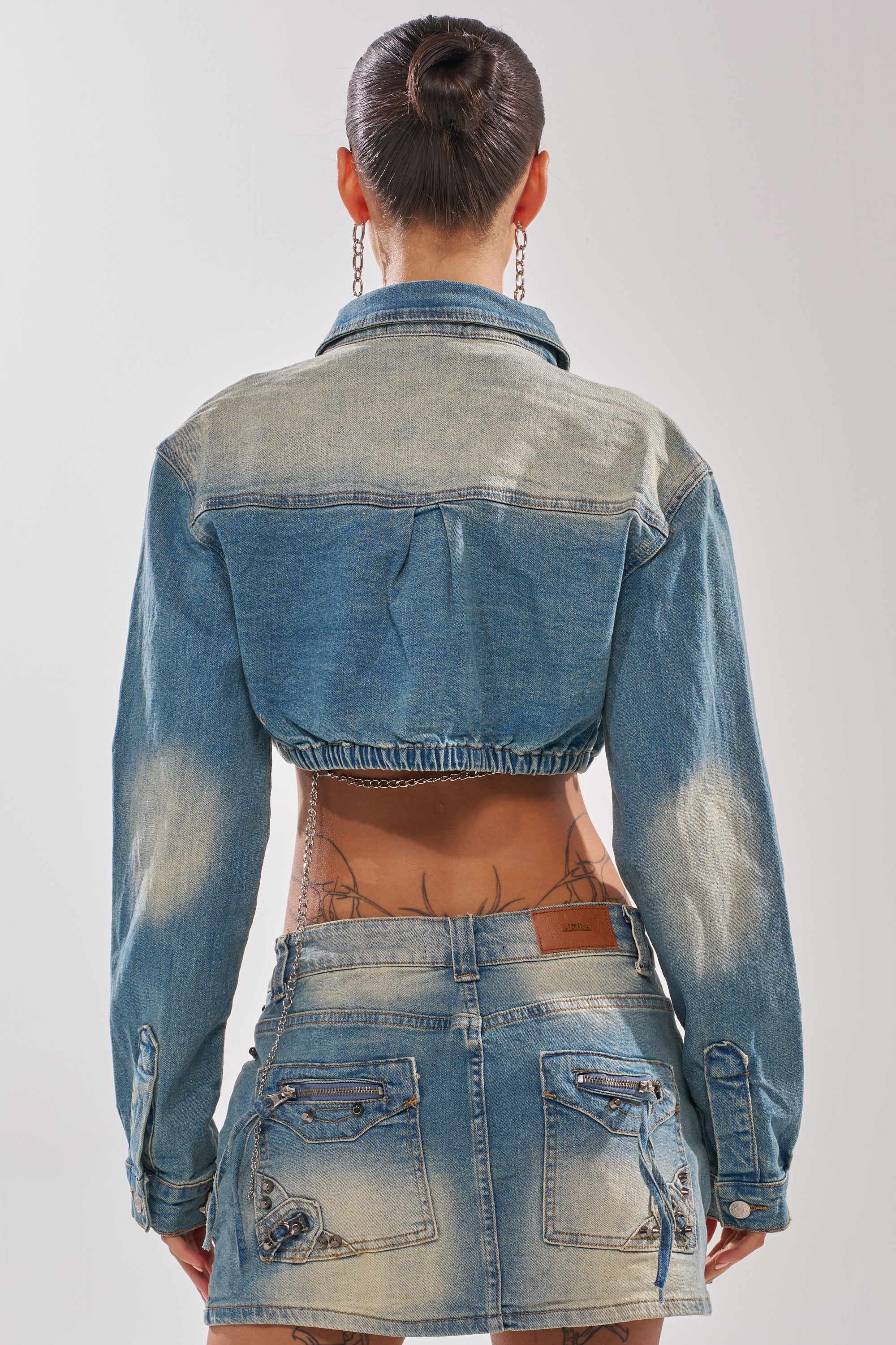 Seen from the back, a person with a neat bun wears the CROWN CROPPED LONG SLEEVE DENIM BUTTON DOWN, styled street-ready with a low-rise distressed mini skirt; tattoos visible on their lower back and arms.