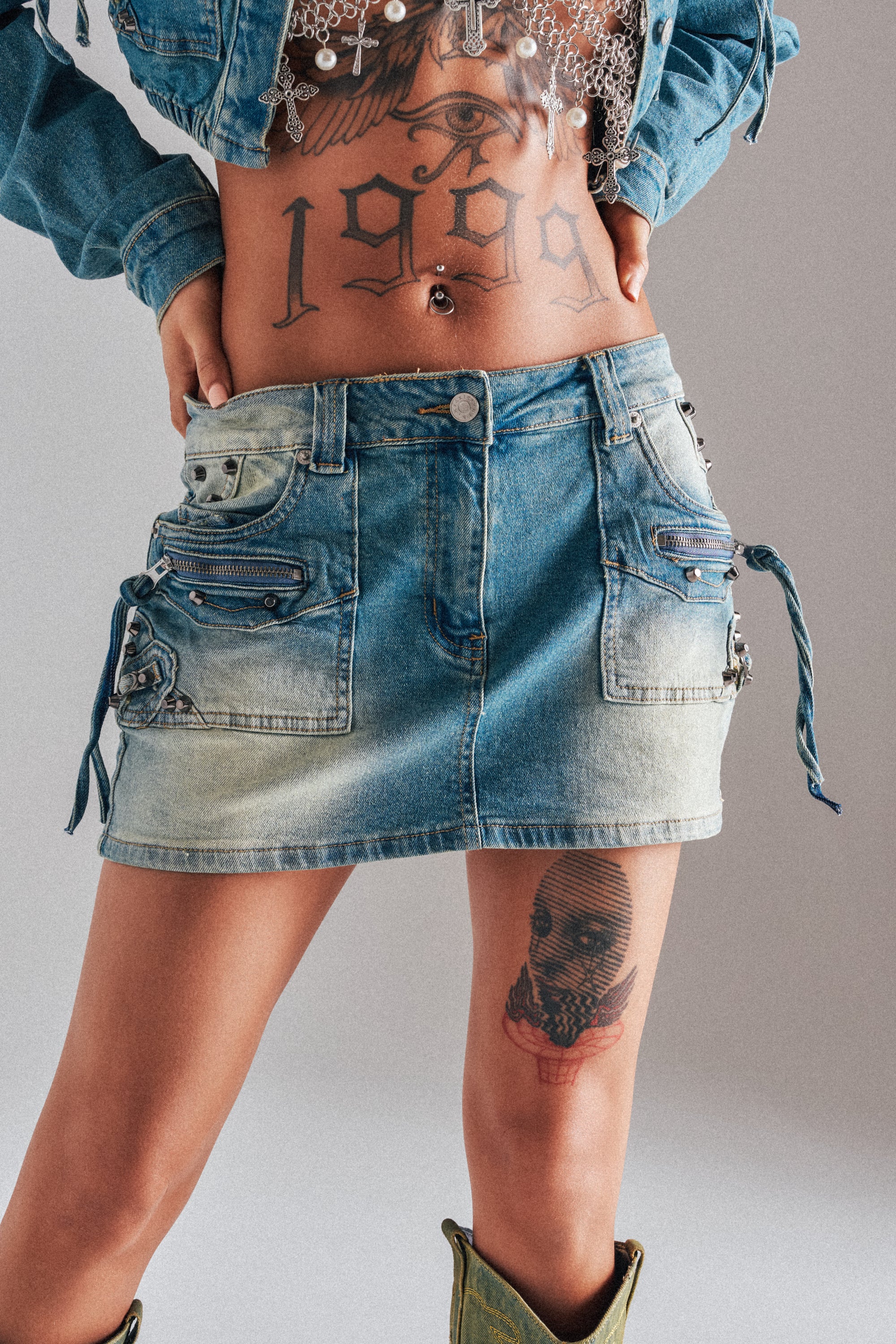 Model wearing the CROWN BELTED DENIM MINI SKIRT and a denim jacket, showing a "1999" tattoo on their stomach and a skull tattoo on their thigh, posed against a neutral background. 0