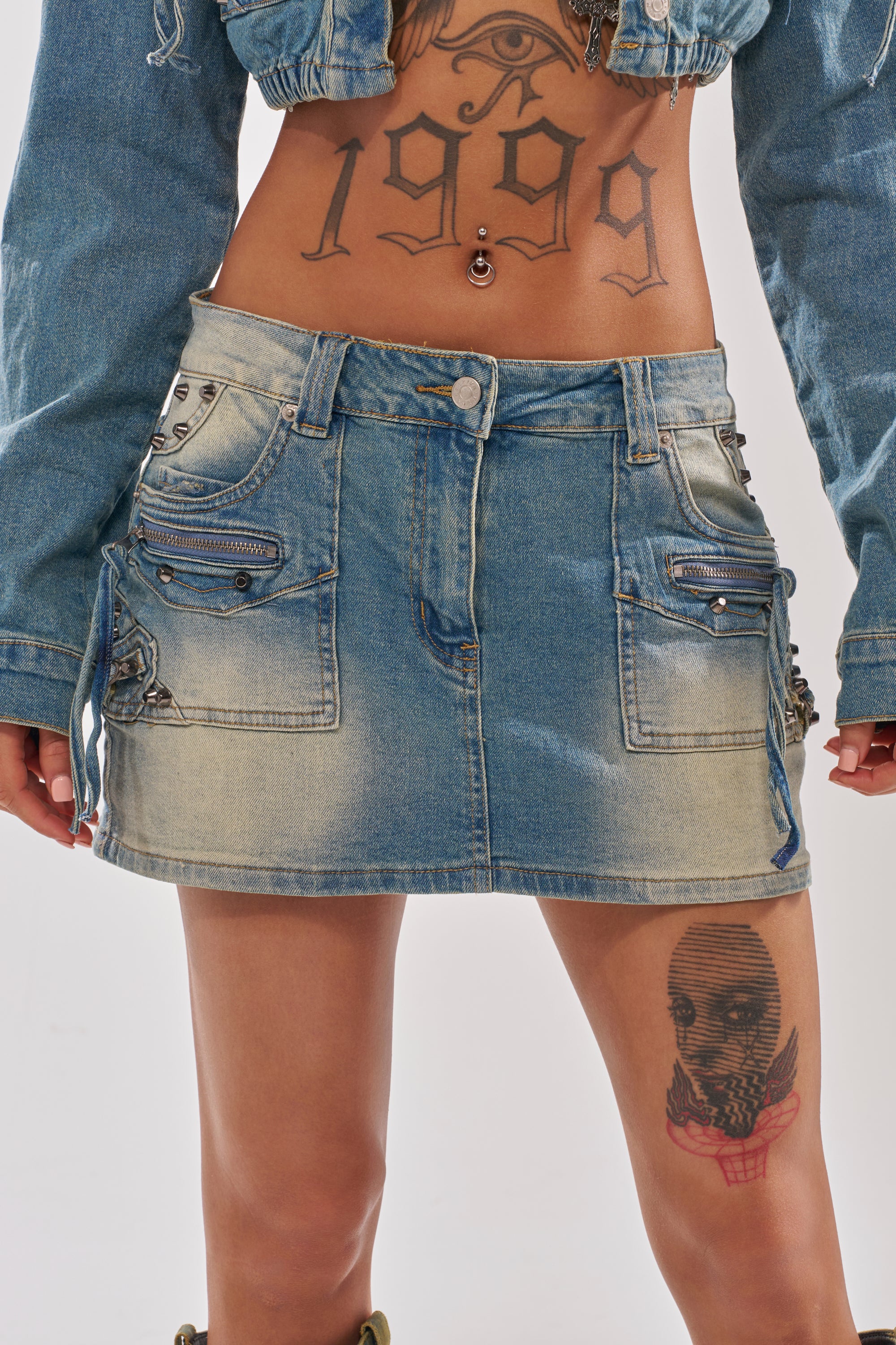 A person wears the CROWN BELTED DENIM MINI SKIRT paired with a matching cropped denim jacket. Tattoos shown are "1999" under an eye on the torso and a skull with a red bow on the thigh. 2