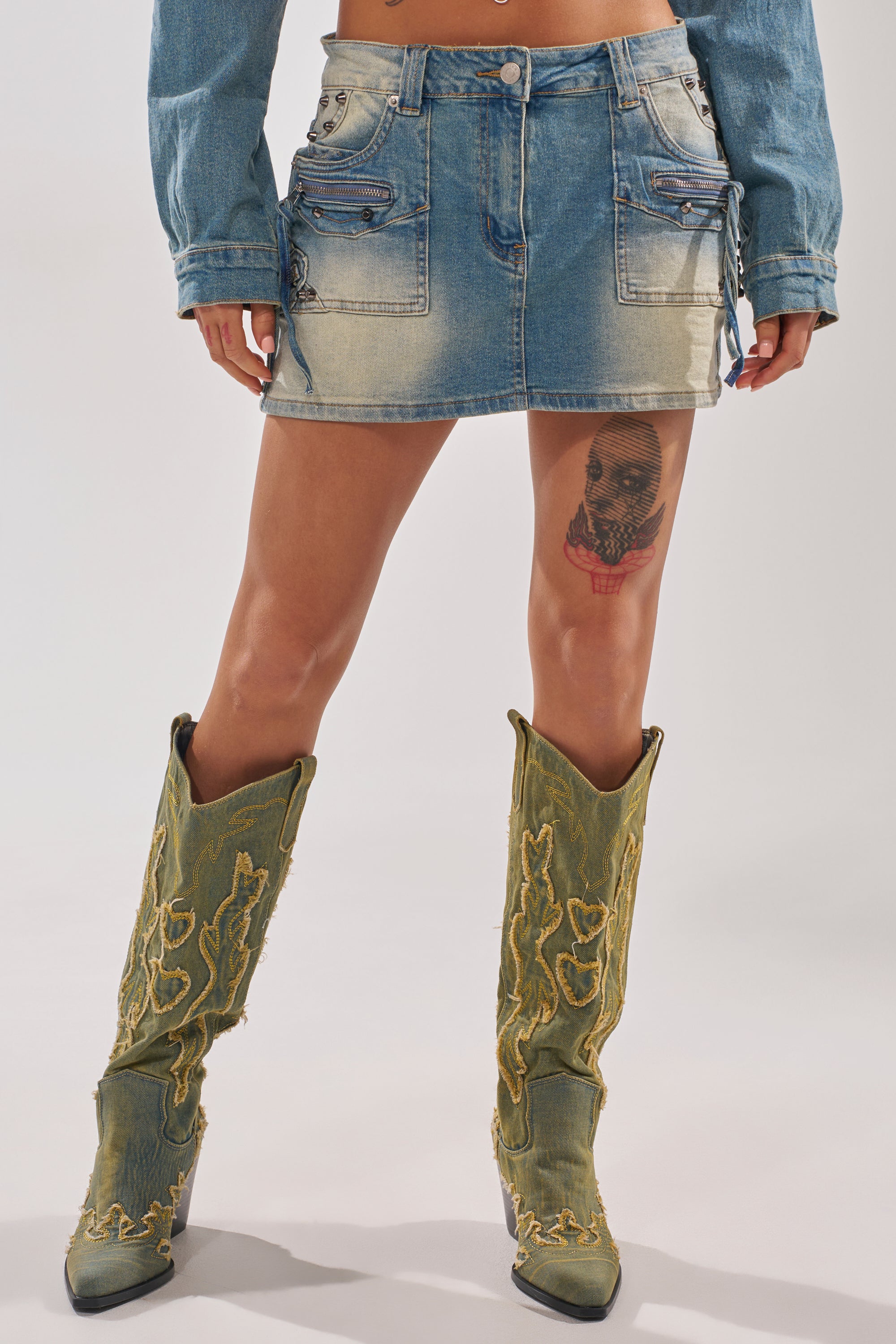A person wears the CROWN BELTED DENIM MINI SKIRT, a long-sleeve denim top, and green cowboy boots with yellow embroidery. Their legs are exposed, revealing a thigh tattoo. 4