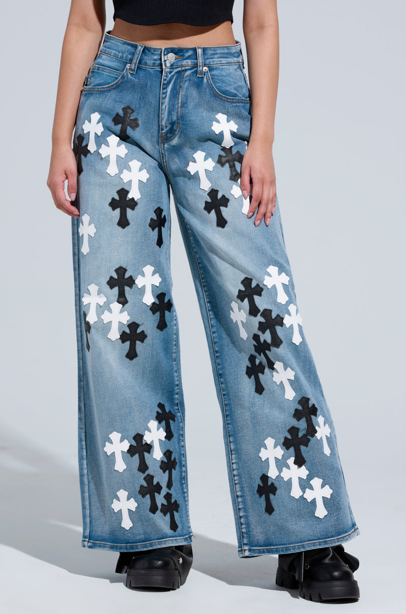 A person wearing the TAKE ME TO CHURCH PATCHWORK DENIM JEAN, featuring black and white cross patterns in a high-rise wide-leg style, paired with black platform shoes. The hands are resting at the sides against a plain light gray background.
