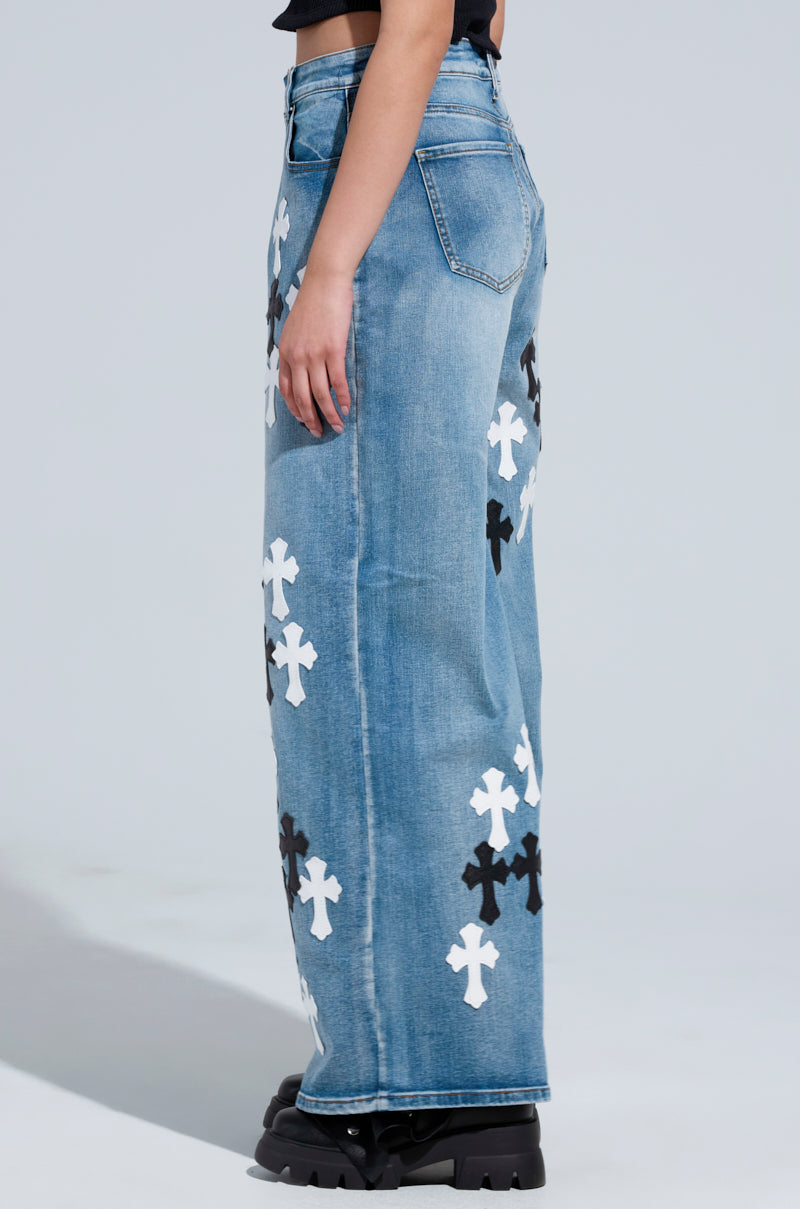 A person wearing the TAKE ME TO CHURCH PATCHWORK DENIM JEAN, featuring high-rise wide-leg design with white and black cross patterns, stands in profile against a light background. The jeans are paired with chunky black shoes, making a bold fashion statement.