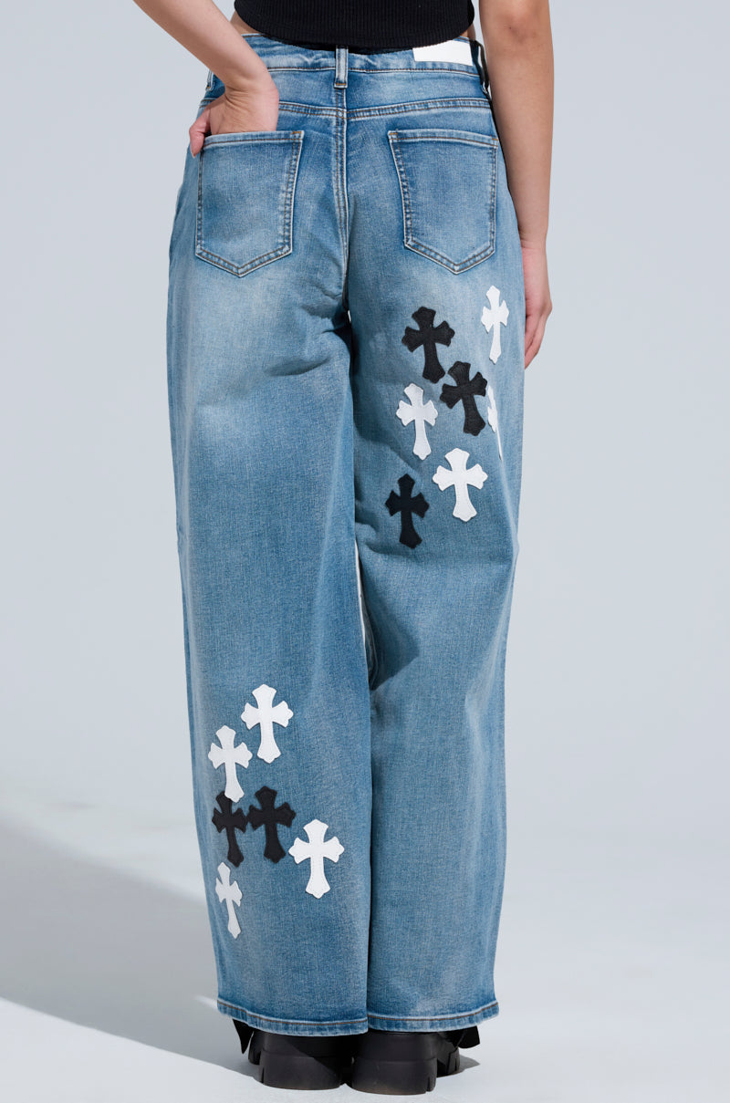 An individual is dressed in the TAKE ME TO CHURCH PATCHWORK DENIM JEAN, which boasts high-rise wide-leg silhouette complemented by striking faux leather cross patches. The design includes back pockets and looks impeccable with sleek black shoes, highlighting the distinctive denim patchwork from an engaging rear view.