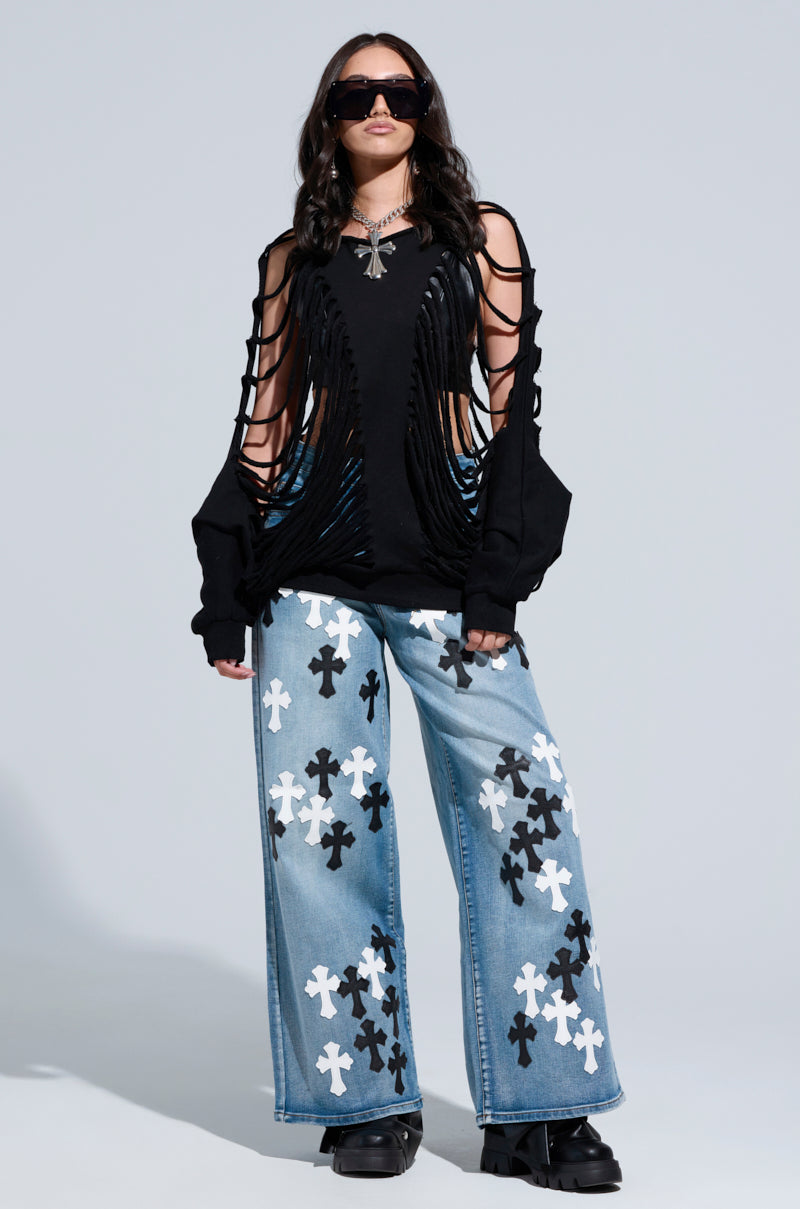 A person in dark sunglasses dons a black, shredded top alongside TAKE ME TO CHURCH PATCHWORK DENIM JEANS featuring black and white cross patterns, exuding confidence. They complete the look with black platform shoes and a statement necklace against a plain backdrop.
