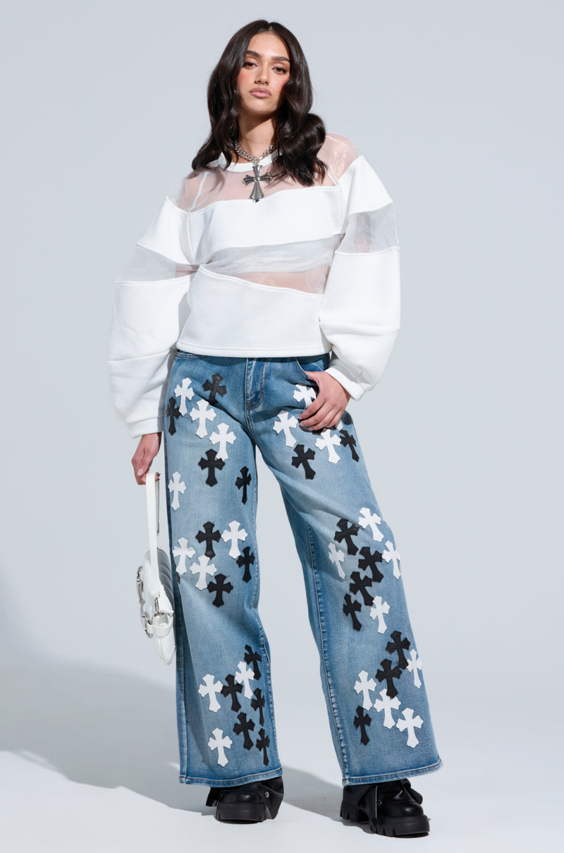 A person stands confidently wearing a white sheer top with wide sleeves, high-rise TAKE ME TO CHURCH PATCHWORK DENIM JEAN with black and white floral patterns, and black shoes. They hold a white purse and have long dark hair against a plain light gray background.