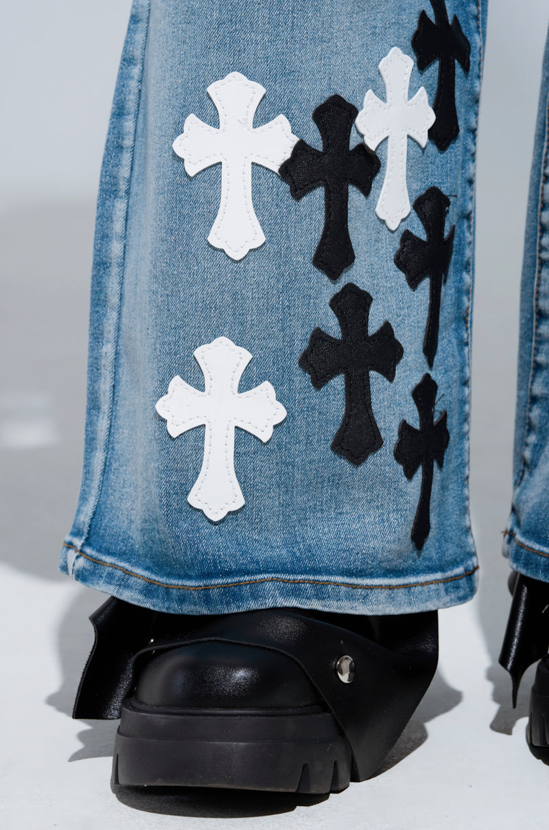 Close-up of the TAKE ME TO CHURCH PATCHWORK DENIM JEAN, featuring white and black faux leather cross patches. The jeans are paired with black shoes, creating a contrasting and stylish look against a plain background.