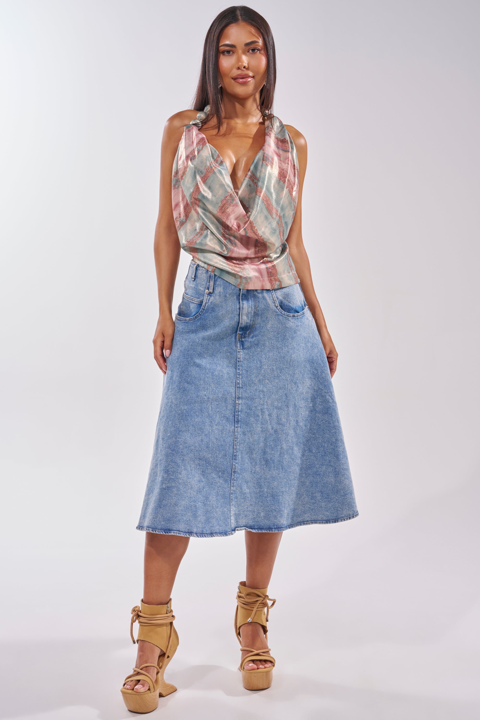 A woman models a sleeveless, draped, multicolored top paired with the BRITNEY MIDI DENIM SKIRT—a high-waisted, A-line fit and flare midi skirt—and tan wedge sandals with ankle ties, capturing effortless streetwear style. 0