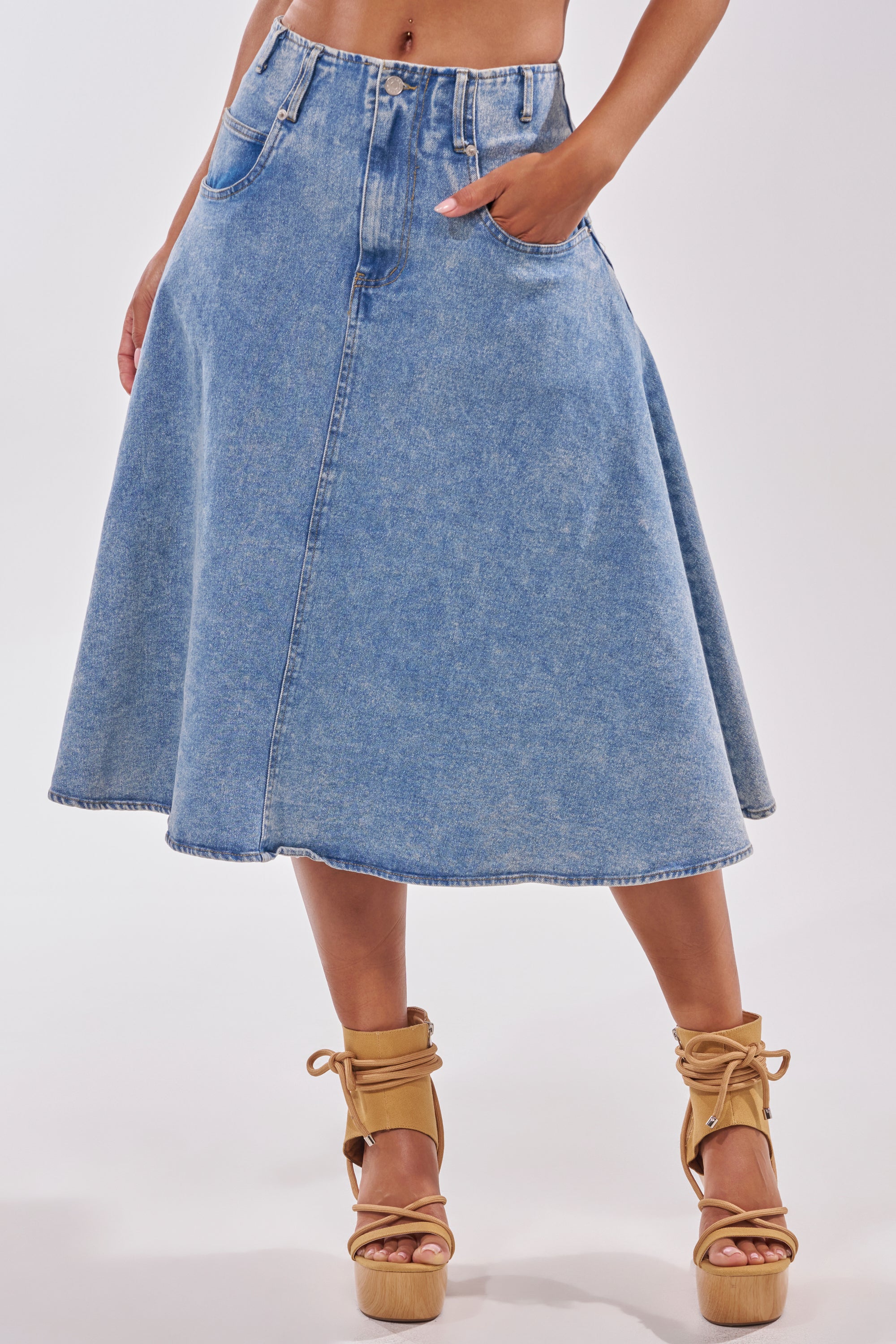 Wearing the BRITNEY MIDI DENIM SKIRT in light blue, high waisted and flared with front pockets, styled with tan platform ankle-strap sandals—this casual streetwear look is completed by posing with one hand in a pocket. 1