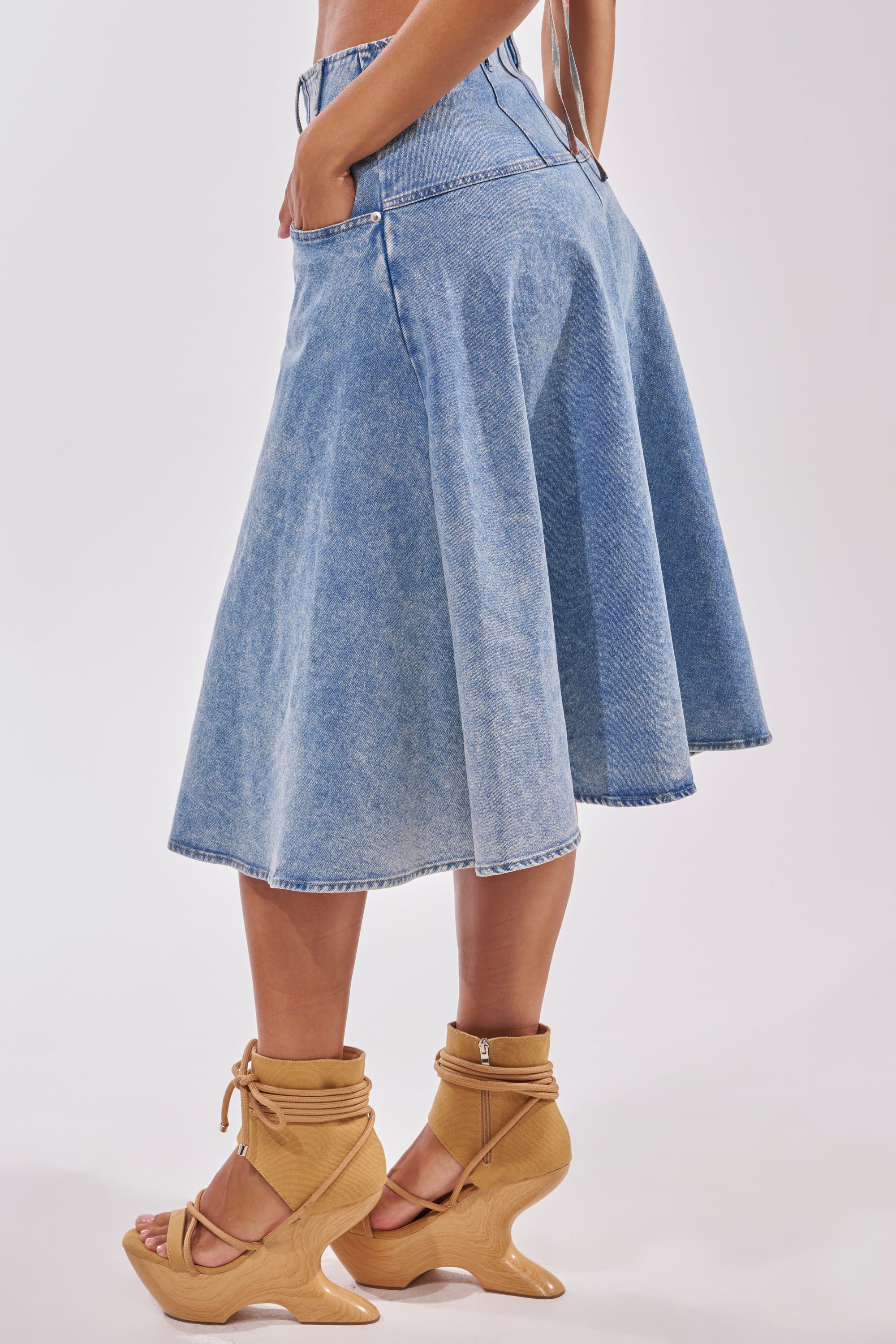 Wearing the BRITNEY MIDI DENIM SKIRT, high-waisted and flared in blue denim, with hands in pockets and tan sculpted-heel sandals, a person showcases casual streetwear style against a light background. 3