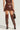 A person is viewed from behind, dressed in the ALL YOURS SIDE LACE UP FAUX CROCODILE MINI SKIRT IN BROWN with a bodycon fit, a coordinating top, and brown over-the-knee boots adorned with decorative patterns and studs. They are carrying a small brown handbag.