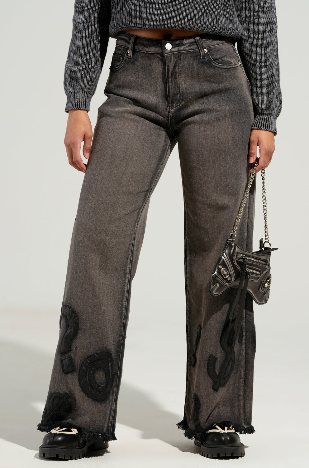 A person dressed in READ MY LIPS PATCHWORK DENIM JEANS with black patches and a gray sweater is holding a small black handbag featuring a chain strap. They are also wearing black platform shoes, and the jeans have a fringed hem at the bottom.