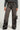 A person dressed in READ MY LIPS PATCHWORK DENIM JEANS with black patches and a gray sweater is holding a small black handbag featuring a chain strap. They are also wearing black platform shoes, and the jeans have a fringed hem at the bottom.
