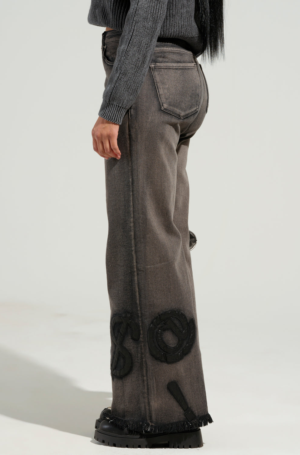 A person wearing a gray sweater and the READ MY LIPS PATCHWORK DENIM JEANS, which feature faded black fabric with fringed hems and embroidered faux leather patches on the wide-leg silhouette. The photo is taken from the side, highlighting the jeans' intricate details.