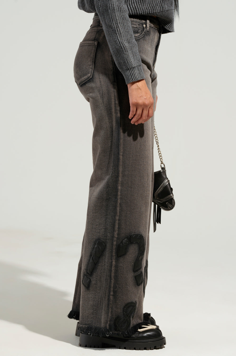 A person is wearing the READ MY LIPS PATCHWORK DENIM JEANS featuring gray tones and a wide-leg silhouette, paired with a cropped sweater. They're carrying a small black handbag with a chain strap, showcasing frayed hems on the jeans, and completing the look with black platform shoes against a light gray background.