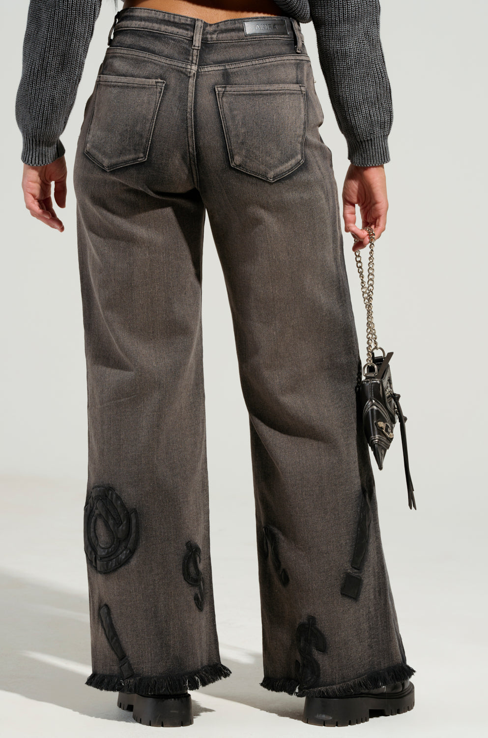 A person wearing a gray sweater and READ MY LIPS PATCHWORK DENIM JEANS, which have a wide-leg silhouette with embroidered faux leather patches, fringed hems, and display dollar signs along with other symbols on the plain background, is holding a small black handbag.