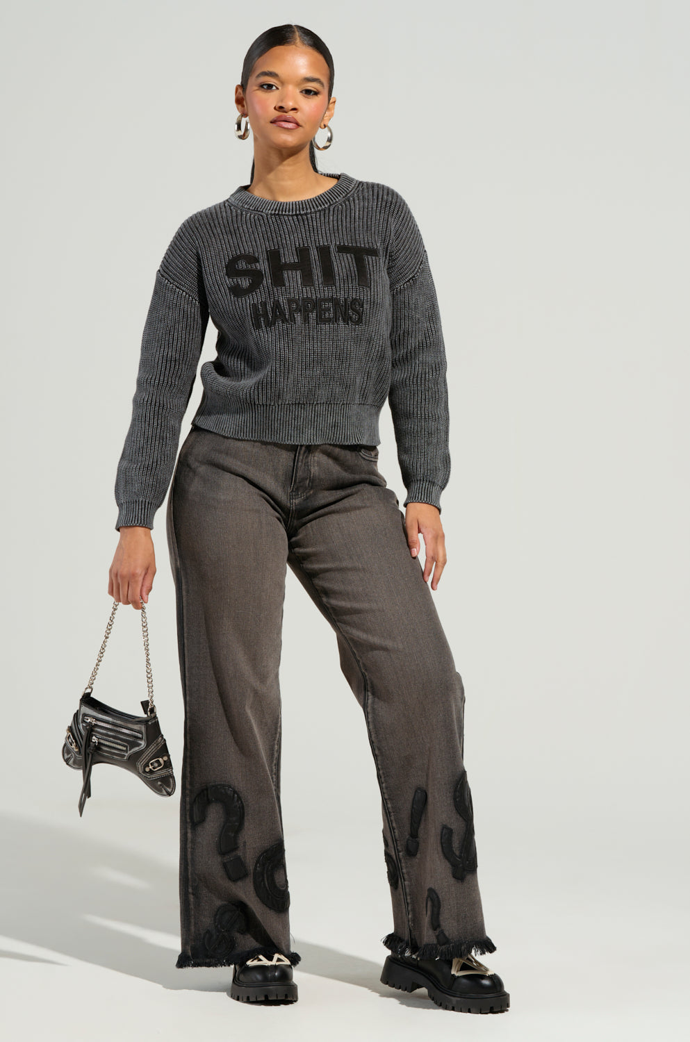 A woman stands confidently in the READ MY LIPS PATCHWORK DENIM JEANS, featuring a wide-leg silhouette, and pairs them with a gray sweater adorned with bold text. She complements her outfit with black shoes and carries a small chain handbag, her hair tied back as she poses against a plain background.